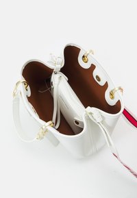 Biała skórzana torebka z otwartą górą, ukazująca brązowe wnętrze oraz zapinaną na zamek kieszeń w środku, z złotymi elementami i czerwono-białym paskiem na ramię.