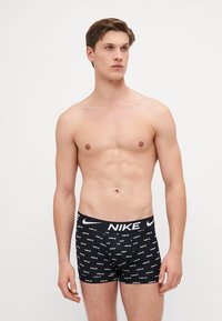 Schwarze Boxer-Shorts aus elastischem Material mit einem sich wiederholenden Muster aus weißen Nike-Logos auf dem Stoff und einem breiten elastischen Bund.