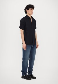 Camicia a maniche corte, con bottoni, in tessuto navy scuro e dal tocco liscio, abbinata a jeans blu aderenti e scarpe nere stringate.