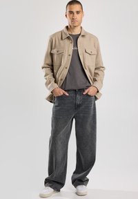 Giacca di flanella beige con due tasche frontali, abbinata a jeans neri sbiaditi a vestibilità ampia e sneaker bianche. Design semplice, look casual.