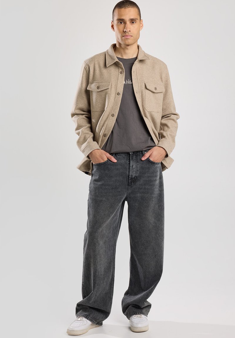 Giacca di flanella beige con due tasche frontali, abbinata a jeans neri sbiaditi a vestibilità ampia e sneaker bianche. Design semplice, look casual.