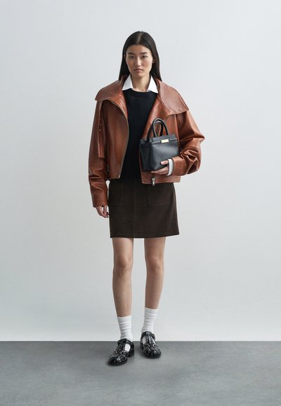 Casaco oversized de pele castanha com colarinho largo, suéter preto, saia castanha, meias brancas e sapatos pretos com adornos. A segurar uma pequena mala preta.