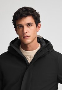 Giacca esterna nera con cappuccio, caratterizzata da una texture liscia e chiusura con zip, indossata sopra un maglione di maglia chiaro.