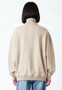 Sweat-shirt beige surdimensionné avec un col montant et des poignets côtelés. Texture douce, design décontracté, et coutures d'épaule tombantes.