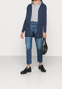Marineblauer Cardigan mit offenem Frontbereich, zwei Taschen, über einem gestreiften Hemd getragen, kombiniert mit blauen Jeans und schwarzen Slippern, hält eine schwarze Handtasche.