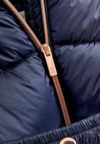 Zbliżenie na granatową pikowaną kurtkę z częściowo otwartym zamkiem błyskawicznym w kolorze różowego złota, posiadającym tłoczony uchwyt w kształcie litery "B".