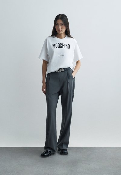 Frau, die mit einer Hand in der Tasche steht, trägt ein weißes Moschino Milano T-Shirt, graue Hose, schwarzen Gürtel und schwarze Schuhe auf einem grauen Boden.