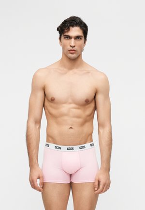 Jaunas raumeningas vyras su tamsiais plaukais dėvi šviesiai rožinius bokserius su baltu dirželiu, ant kurio parašyta „GCDS“, stovintis priešais paprastą baltą fone.