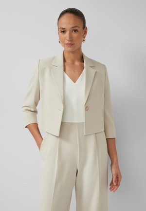 Ženska z lasmi, počešenimi nazaj, ki nosi zlata obročka, krajšeno bež jakno, belo V-izrez majico in bež hlače z visokim pasom, roke v žepih.