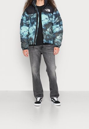 Personne portant une doudoune tie-dye bleu et noir de The North Face, un t-shirt noir, un jean gris délavé et des baskets noires et blanches, debout sur un sol gris.