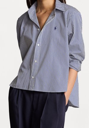 Femme portant une chemise boutonnée à rayures verticales bleu marine et blanches avec les manches retroussées et un pantalon bleu marine foncé, la main dans la poche.