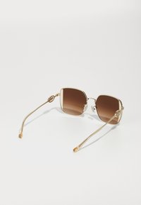 Lunettes de soleil avec accents dorés, dotées de verres bruns dégradés en forme carrée et de branches métalliques fines avec des détails circulaires décoratifs.