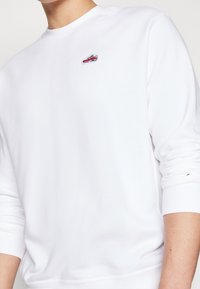 Hombre vestido con un sudadera blanca de manga larga con un pequeño parche de zapatilla roja y negra en el lado izquierdo del pecho.