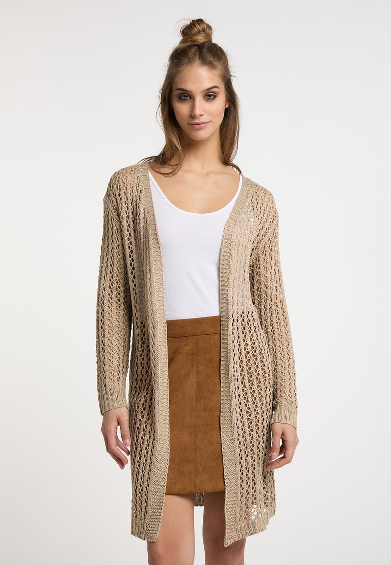Cardigan beige lavorato a maglia con un design a trama aperta, maniche lunghe e vestibilità ampia. Abbinato a un top bianco e una gonna in suede marrone.
