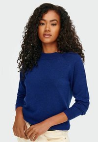 Sinine kootud sveller ümmarguse kaeluse, soonikkoes varrukate ja äärisega; pehme tekstuur; vormiline disain; koos heledate pükstega.