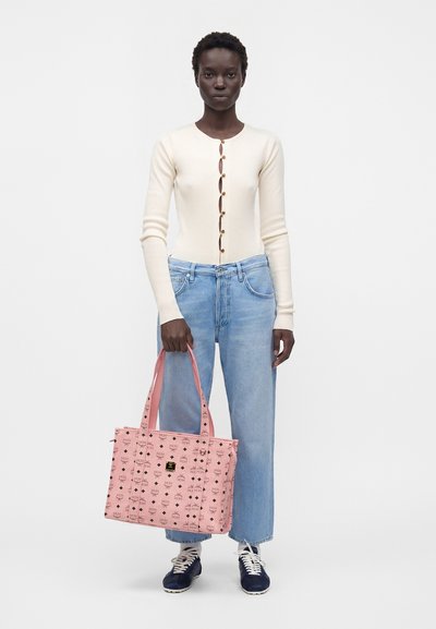 Femme portant un haut crème à côtes manches longues et un jean bleu clair, tenant un sac fourre-tout rose à motifs, debout sur un fond blanc.