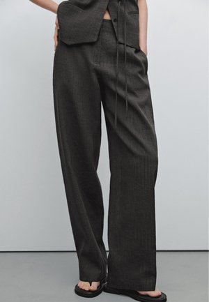 Modèle portant un pantalon large noir à fines rayures et un gilet sans manches assorti, associé à des sandales noires sur un fond uni.