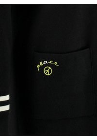 Czarny element garderoby z prostokątną kieszenią ozdobioną zielonym haftem z napisem „peace” nad symbolem pokoju. Białe paski przy krawędzi.