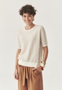 Suéter texturizado color crema con un patrón ondulado, cuello y puños acanalados. Combinado con pantalones de pierna ancha marrones y pulseras gruesas.