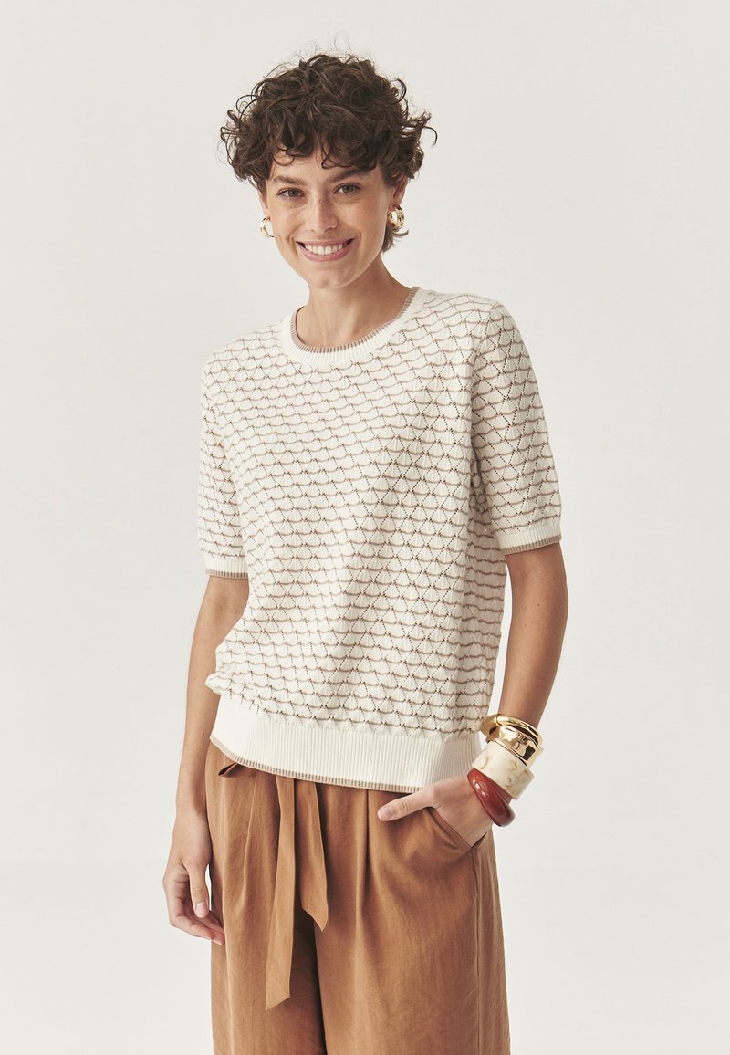Suéter texturizado color crema con un patrón ondulado, cuello y puños acanalados. Combinado con pantalones de pierna ancha marrones y pulseras gruesas.