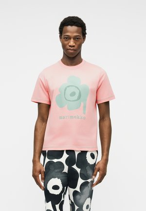 Férfi rózsaszín pólóban, melyen zöld virágmotívum és felirat látható, fekete-fehér virágmintás nadrággal, fehér háttér előtt áll.