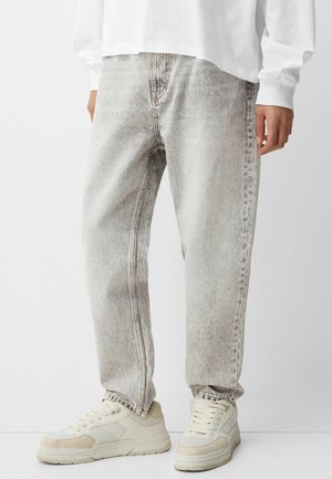 Personne portant un jean délavé gris clair à coupe décontractée et des baskets épaisses blanc cassé, debout devant un fond clair uni.