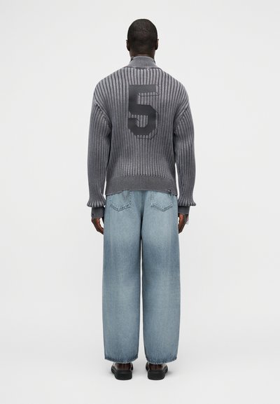 Άνδρας που φοράει γκρι πλεκτό πουλόβερ με μεγάλο μαύρο αριθμό 5 στην πλάτη και ανοιχτόχρωμο τζιν με φαρδιά πόδια, στέκεται με την πλάτη προς τα εμάς.