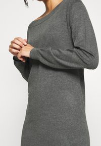 Femme portant une robe en tricot gris foncé à manches longues, les mains jointes près de la poitrine, sur un fond clair uni.