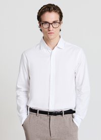 Weißes, langärmliges Hemd mit Knopfleiste und spitzem Kragen, kombiniert mit hellgrauer Hose und schwarzem Gürtel. Glatte Textur; figurbetontes Design.