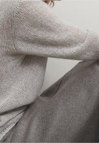 Personne portant un pull léger en maille et un pantalon gris, assise avec les bras posés sur les genoux pliés sur un fond uni.