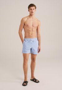 Kék és fehér csíkos fürdőnadrág, elasztikus derékkal és húzózsinórral, oldalsó zsebekkel és laza szabással. Fekete papucs viselve.