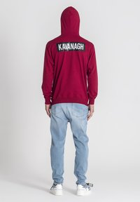 Sudadera con capucha color marrón burdeos con un logo negro "KAVANAGH" en la parte posterior. Combinada con jeans de color azul claro y zapatillas blancas con detalles.
