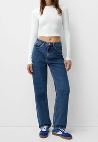 Vit långärmad kort topp, högmidjade blå denimjeans med rak ben, bärs med blå och vita sneakers på en neutral bakgrund.