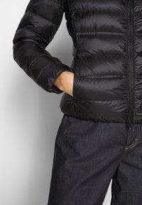 Sort pufferjakke med et quiltet design, lynlås foran og elastiske manchetter. Sammen med mørke denimbukser med et subtilt mønster.