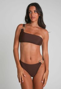 Brun bikini med en axel-design, med guldfärgade detaljer på remmen och sidan av trosorna. Slät textur, minimal täckning.