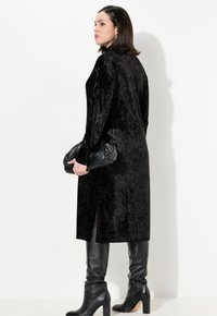 Mantello lungo in velluto nero con finitura testurizzata, caratterizzato da fessure laterali, indossato sopra stivali neri in pelle con tacco alto, accessoriato con una pochette nera.