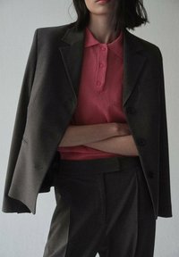 Blazer grigio scuro sopra una polo rosa con tre bottoni. Il blazer ha un risvolto classico e spalle strutturate. Tessuto dalla texture liscia.