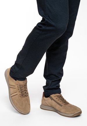 Beine in dunkler Hose und beige Wildleder-Sneakern mit Schnürsenkeln und seitlichem Reißverschluss, ein Fuß leicht angehoben vor weißem Hintergrund.