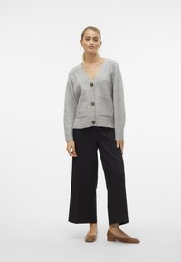 Cardigan en maille grise avec un col en V, agrémenté de grands boutons marron et de deux poches avant, associé à un pantalon noir à jambes larges et des chaussures plates marron.