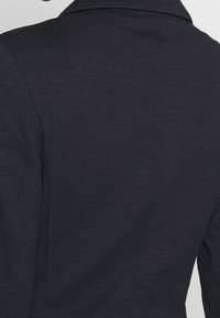 Veste gris foncé texturée avec un tissu doux à côtes. Le col est replié, révélant des coutures lisses et une coupe ajustée à l'arrière.