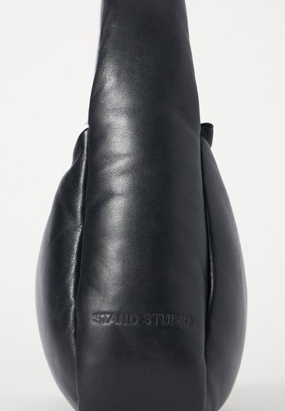 Μαύρη δερμάτινη τσάντα με στρογγυλό σχήμα και ομαλή υφή. Διαθέτει ανάγλυφο λογότυπο "STAND STUDIO" και μινιμαλιστικό σχέδιο.