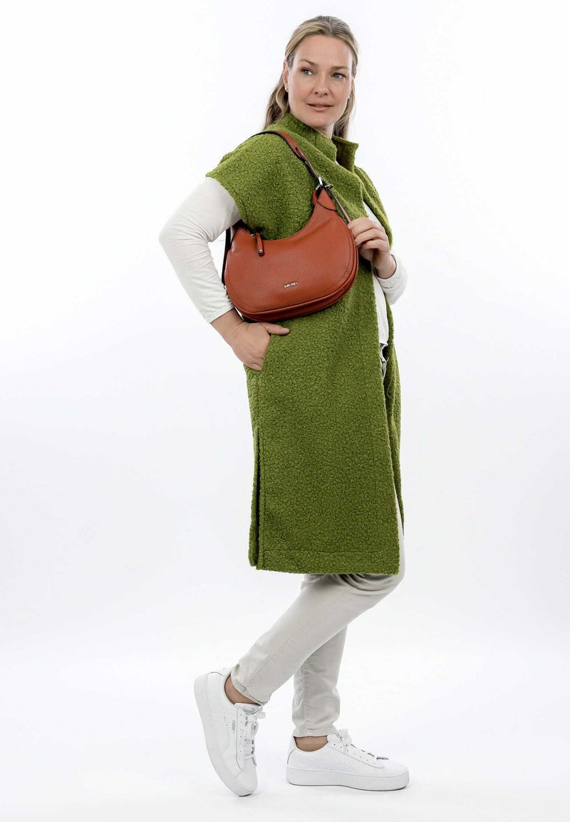 Sac à main en cuir marron avec une forme courbée, surface texturée, et une courte bandoulière, porté avec un long gilet en polaire vert et un pantalon blanc.