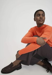 Helles orangefarbene, gerippte Strickjacke mit hohem Kragen, kombiniert mit lockeren, dunklen Hosen und braunen Loafers mit Fransen-Detail.