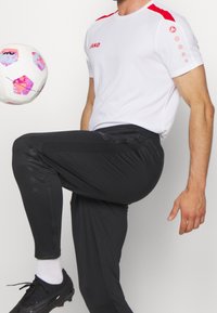 Man i vit och röd sporttröja och svarta byxor som sparkar en vit fotboll med rosa och lila mönster inomhus.