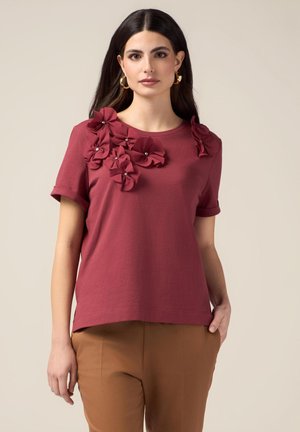 La camiseta de color burdeos presenta flores de tela tridimensionales con acentos de perlas en un hombro; material de algodón suave con mangas cortas.