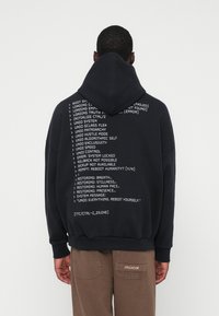 Άνδρας που φοράει μαύρη κουκούλα με λευκό κείμενο τεχνολογικού στυλ στην πλάτη, που στέκεται μπροστά σε απλή ανοιχτόχρωμη background, με τα χέρια του στα πλάγια.