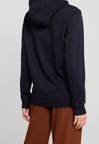 Námořnická mikina vyrobená z bavlněné směsi, která má klokankovou kapsu a žebrované manžety, spárovaná s hnědými kalhotami širokého střihu.