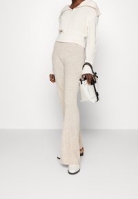 Weiße gerippte, cropped Kapuzenjacke mit einem großen Kragen, kombiniert mit beigen, ausgestellten Hosen, die eine subtile Textur aufweisen. Eine kleine weiße Handtasche wird gehalten.