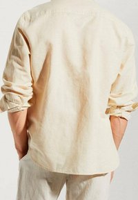 Camisa de manga larga beige hecha de tejido ligero, con dobladillo curvado y mangas recogidas, textura suave con costuras mínimas.