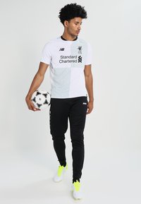 Witte en grijze Liverpool FC-tenue met zwart logo, gecombineerd met zwarte broek en witte voetbalschoenen met neonaccenten, terwijl hij een voetbal vasthoudt.